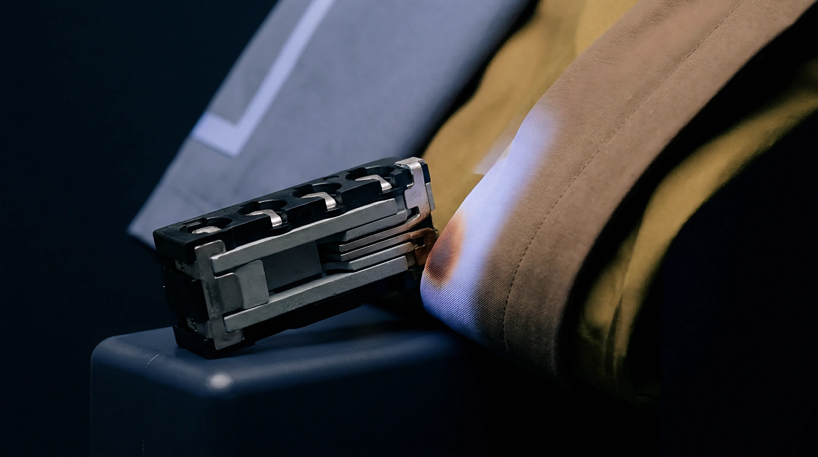 A metal and polymer battery cell interconnect rests beside a scorched textile sample during thermal testing.