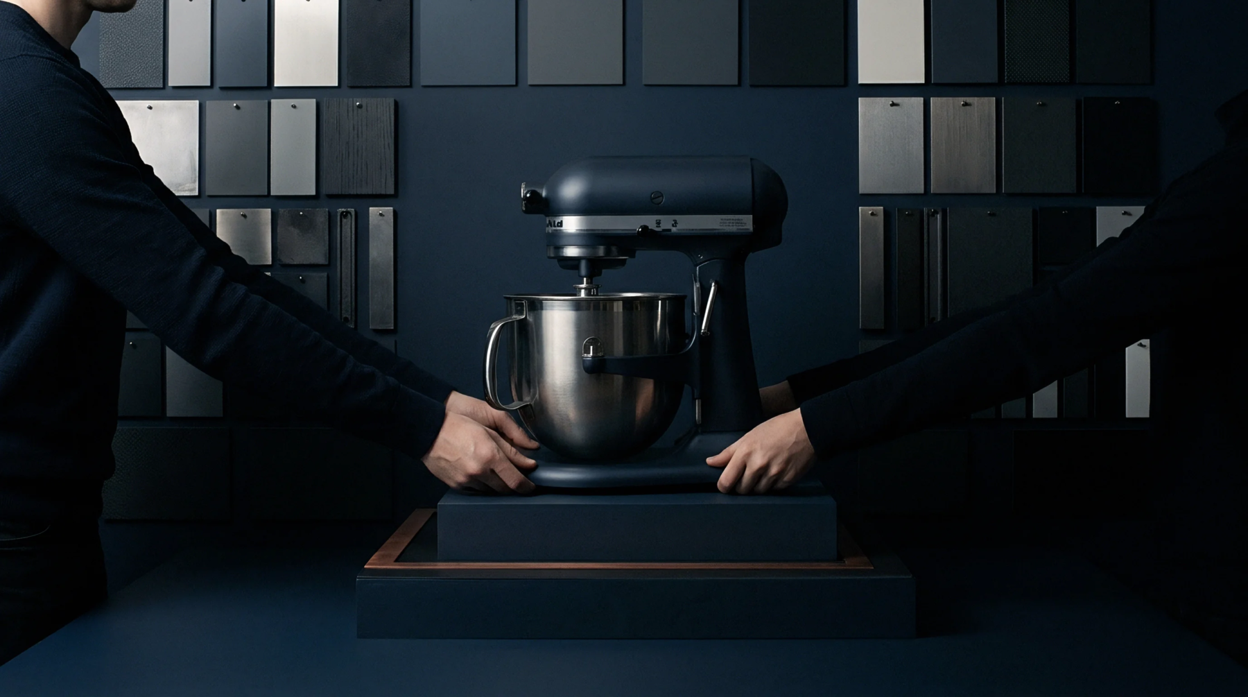 A dark metallic hardware assembly sits on a display platform flanked by human hands before a wall of material sample plates.