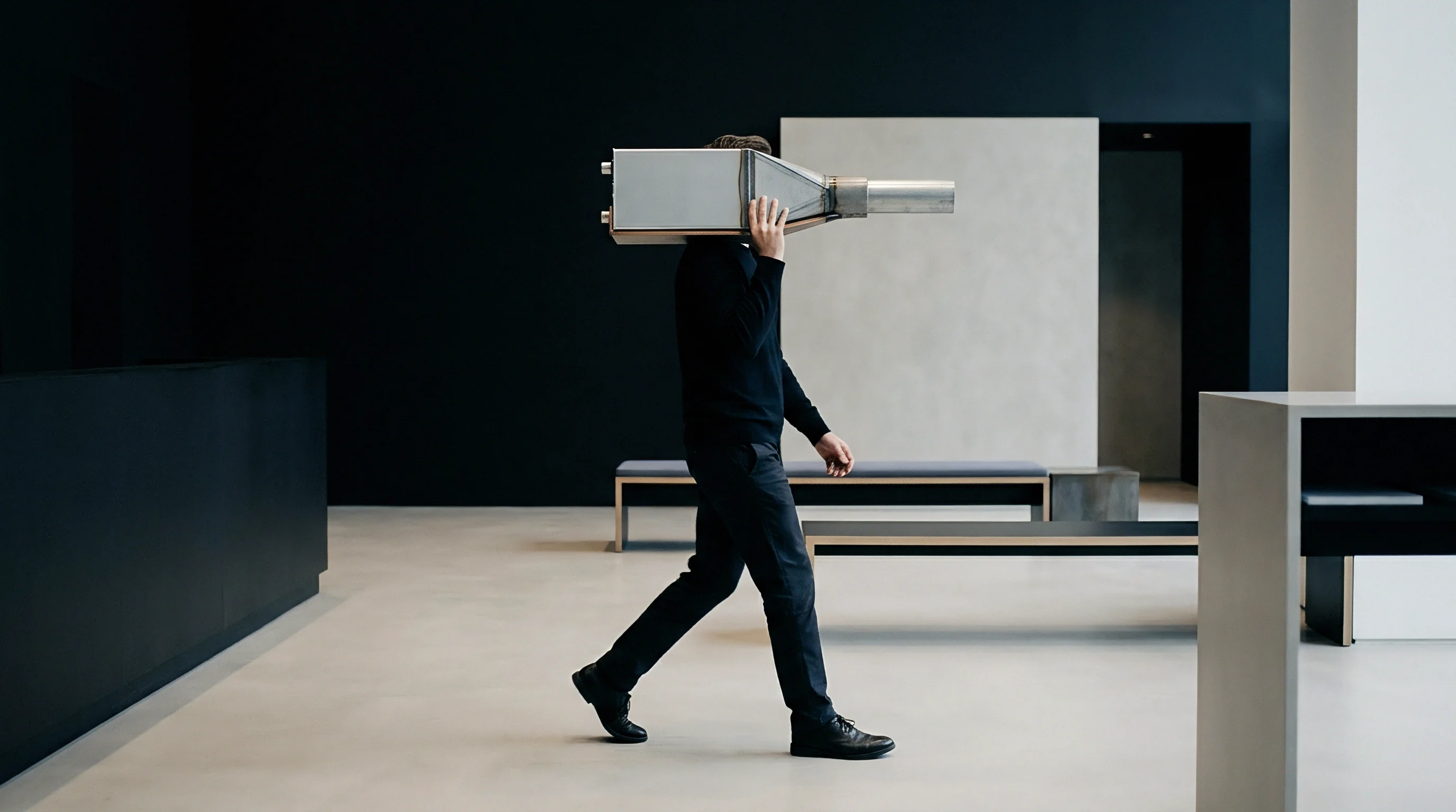 A technician carries a bulky stainless steel hardware assembly across a minimalist industrial production facility floor.