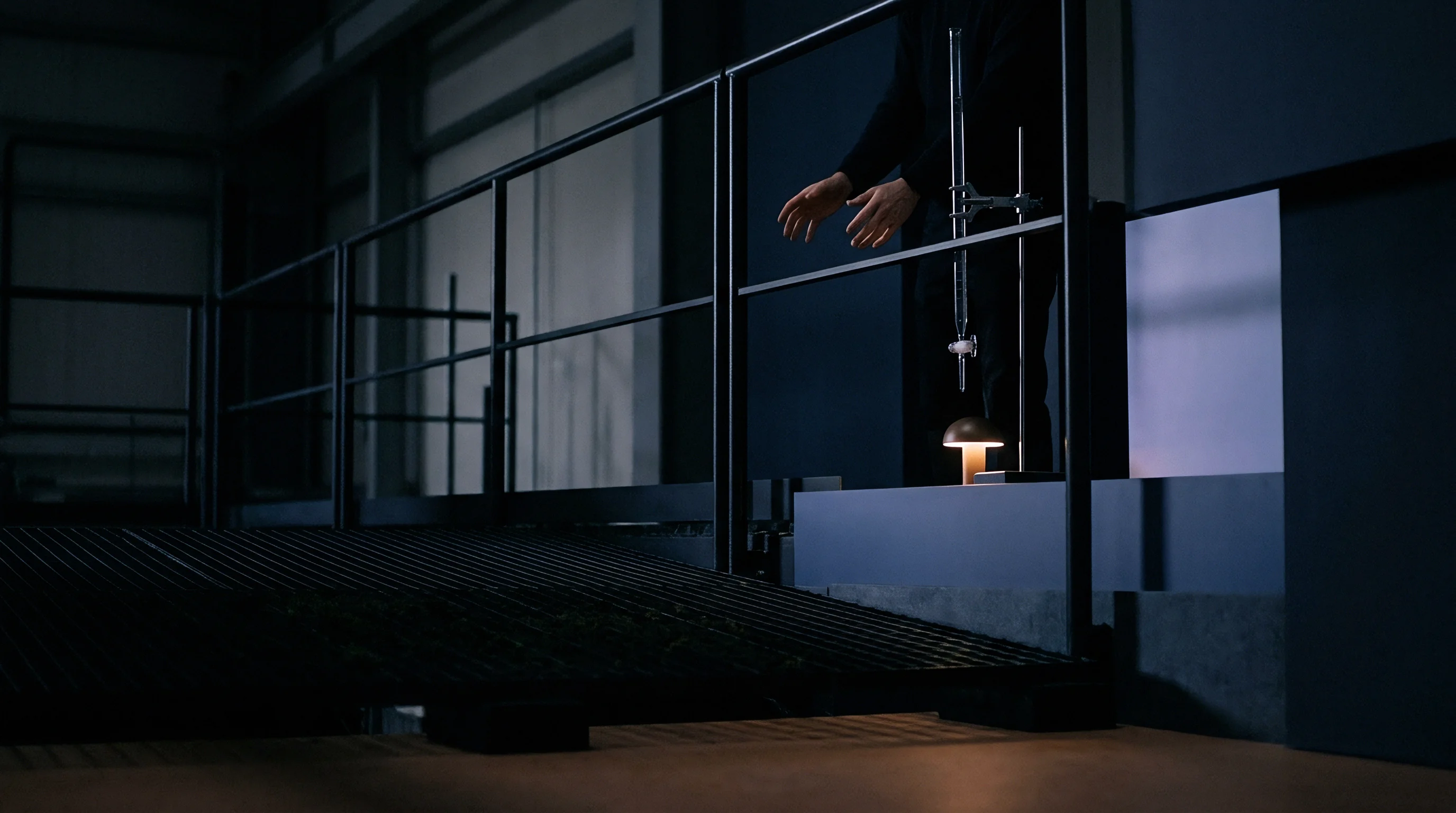A laboratory technician stands behind metal railings beside a titration column mounted over a small glowing lamp in a dark facility.