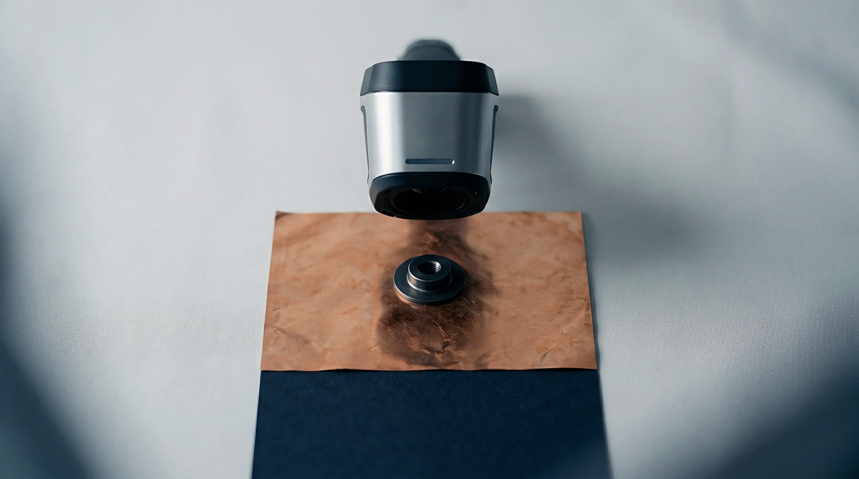A handheld optical measurement tool hovers above a discolored copper foil sample fixed on a dark testing plate.